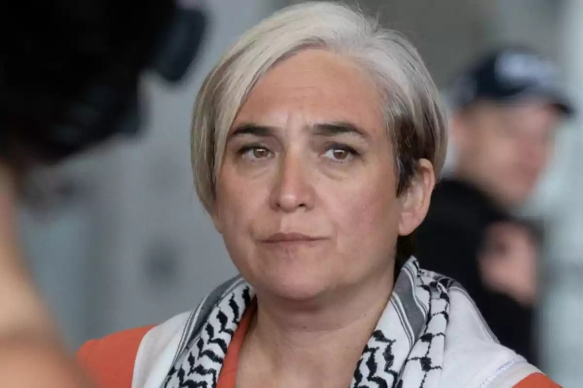 Mujer de cabello corto y canoso con expresión seria, usando una bufanda con estampado blanco y negro y una prenda naranja, en un entorno interior desenfocado. Mujer de cabello corto y canoso con expresión seria, usando una bufanda con estampado blanco y negro y una prenda naranja, en un entorno interior desenfocado.