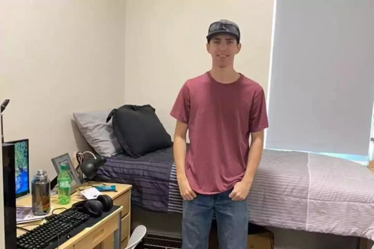 Joven con camiseta roja y gorra de pie junto a una cama en una habitación ordenada con escritorio y computadora