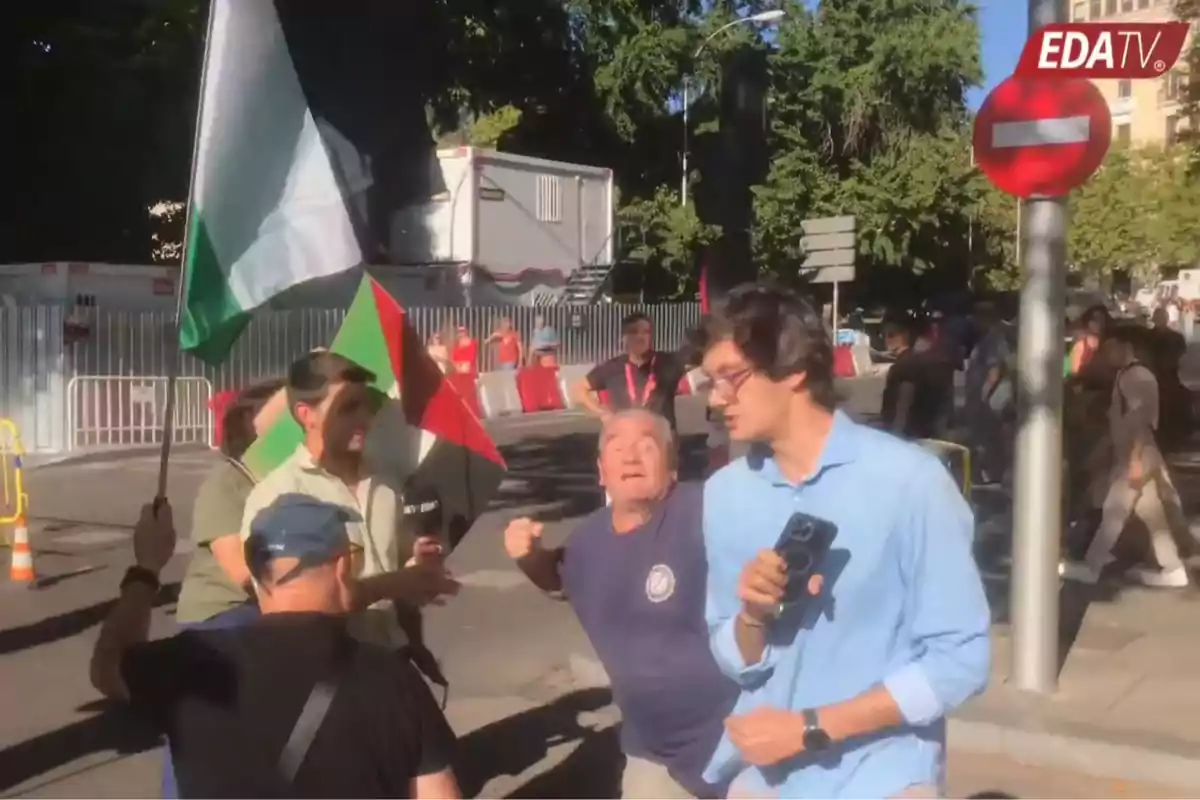 Un grupo de personas discute en la calle durante una manifestación mientras un reportero sostiene un micrófono y varias banderas ondean en el fondo