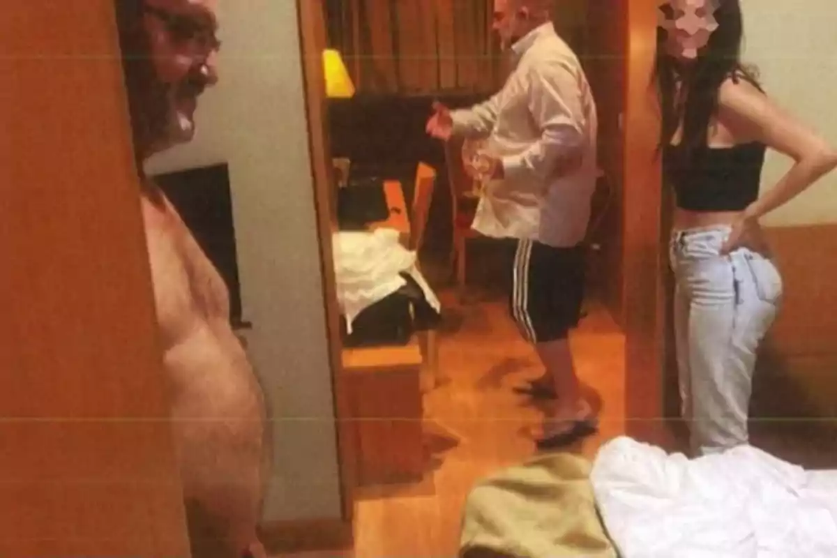 Tres personas en una habitación de hotel, una de ellas sin camisa, otra con camisa y pantalones cortos, y una mujer con el rostro pixelado de pie junto a la cama desordenada