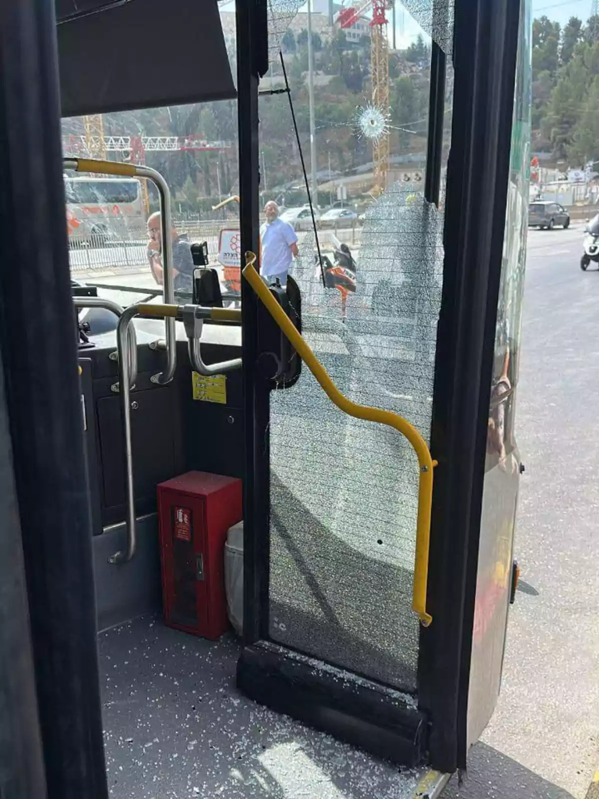 Puerta de autobús con el vidrio roto y un agujero de impacto, fragmentos de vidrio en el suelo y personas observando desde el exterior Puerta de autobús con el vidrio roto y un agujero de impacto, fragmentos de vidrio en el suelo y personas observando desde el exterior
