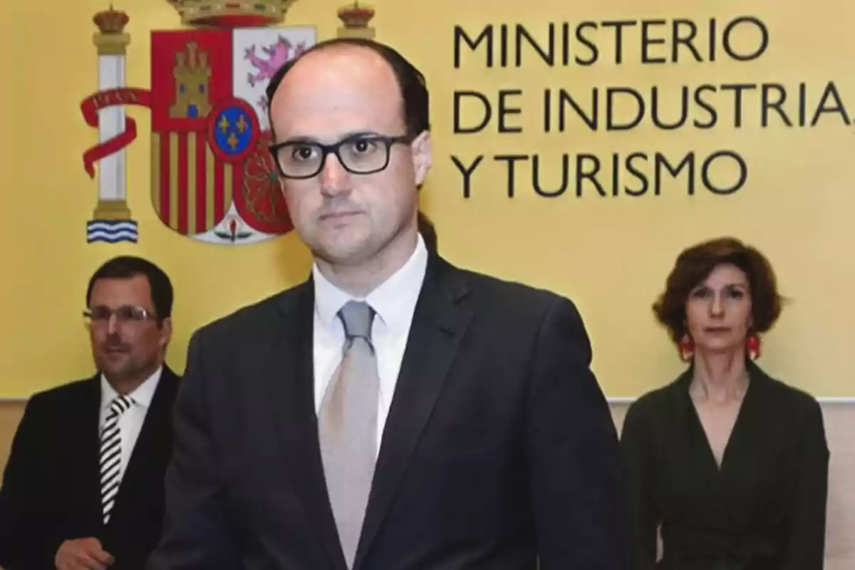 Hombre con gafas y traje frente a un fondo amarillo con el escudo de España y el texto Ministerio de Industria y Turismo acompañado por dos personas en segundo plano Hombre con gafas y traje frente a un fondo amarillo con el escudo de España y el texto Ministerio de Industria y Turismo acompañado por dos personas en segundo plano