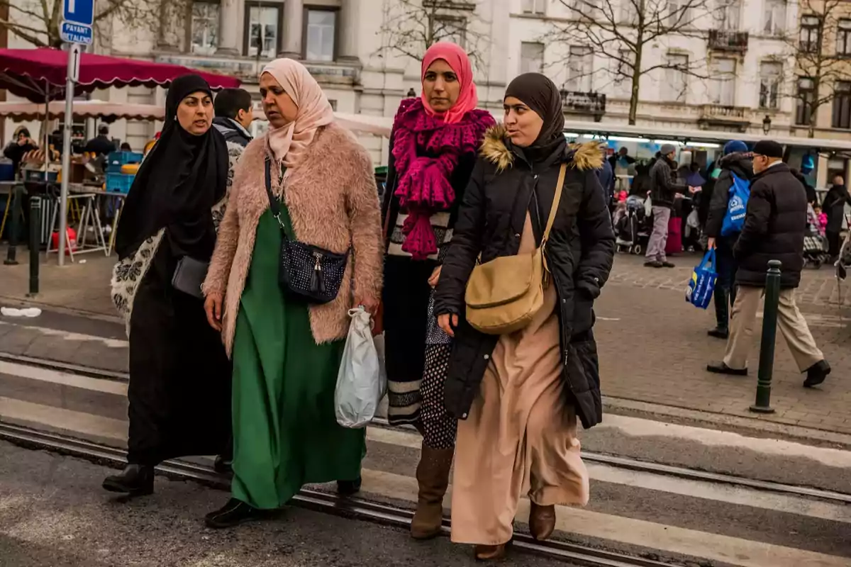 Cuatro mujeres con hiyab caminan juntas por una calle concurrida en una ciudad, al fondo se ven puestos de mercado y varias personas
