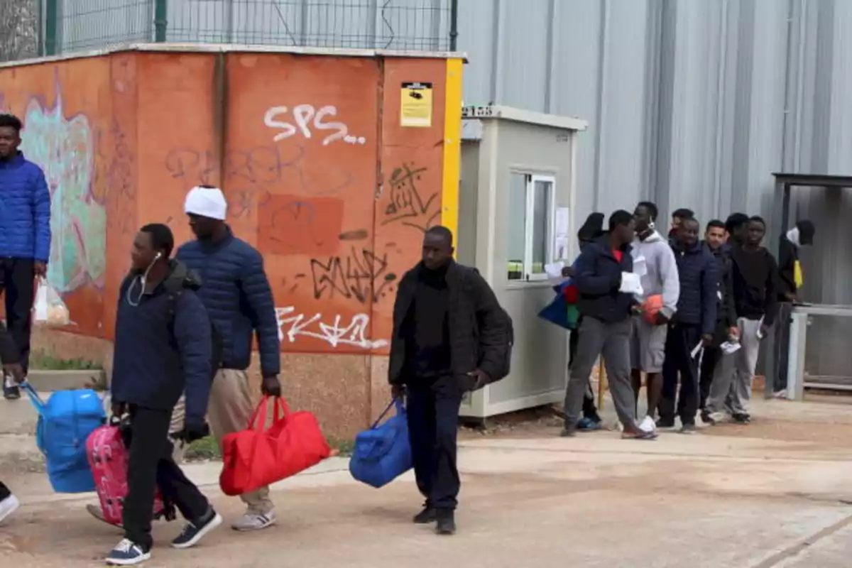 Un grupo de personas camina y hace fila con maletas y bolsas en la mano frente a un edificio con paredes grafiteadas. Un grupo de personas camina y hace fila con maletas y bolsas en la mano frente a un edificio con paredes grafiteadas.