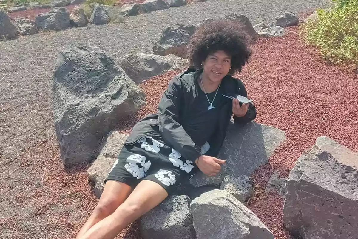 Persona recostada sobre unas rocas en un entorno al aire libre con suelo de grava rojiza y vegetación al fondo, usando ropa oscura y sosteniendo un teléfono móvil mientras sonríe a la cámara