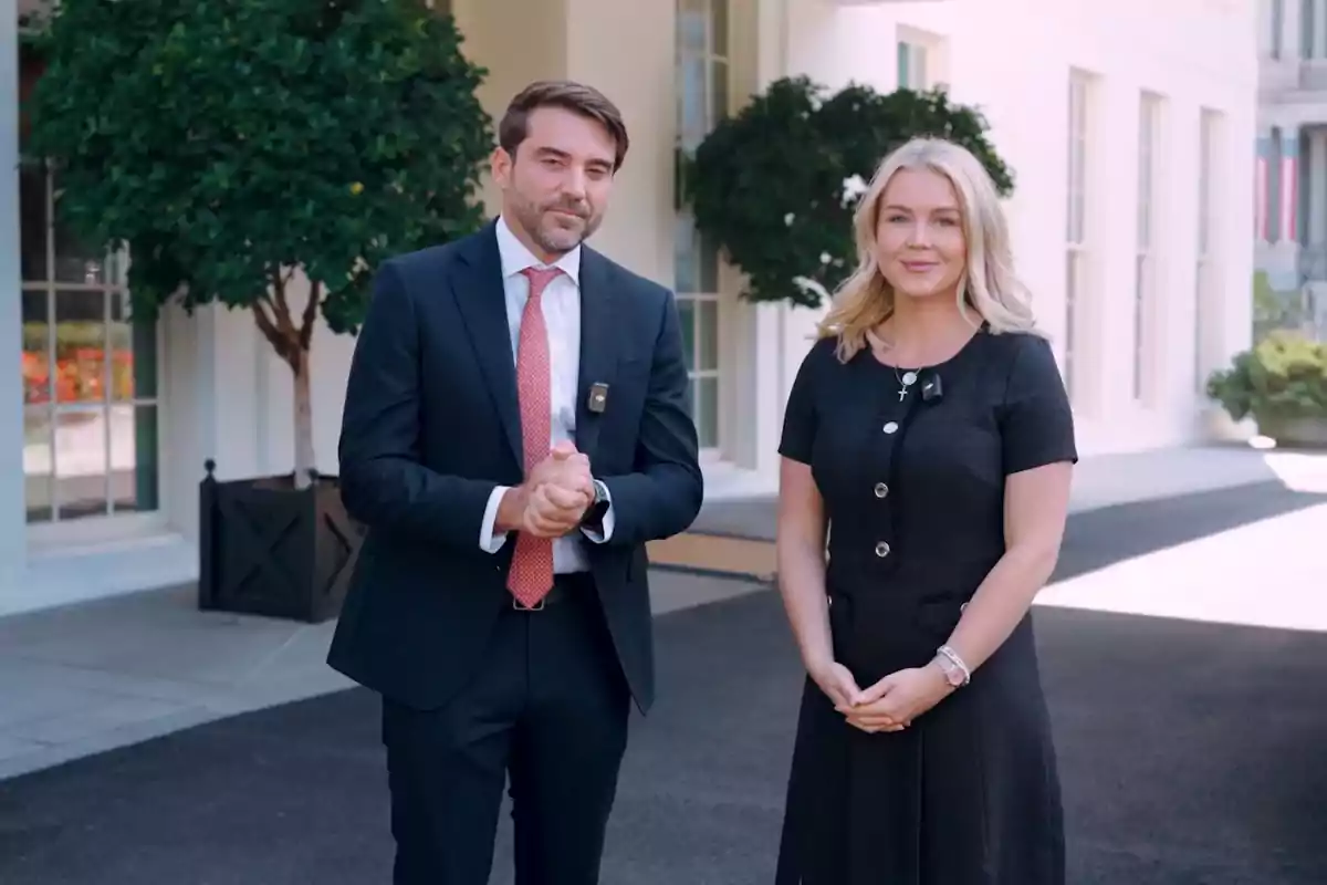 Dos personas de pie al aire libre frente a un edificio elegante con grandes ventanas y árboles en macetas, ambos vestidos de manera formal y sonriendo a la cámara.