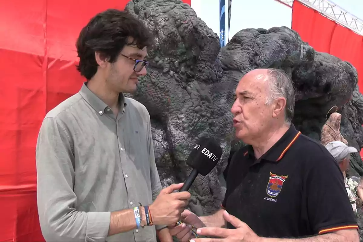 Un joven con gafas sostiene un micrófono mientras entrevista a un hombre mayor con camiseta negra frente a un fondo de rocas y telas rojas Un joven con gafas sostiene un micrófono mientras entrevista a un hombre mayor con camiseta negra frente a un fondo de rocas y telas rojas