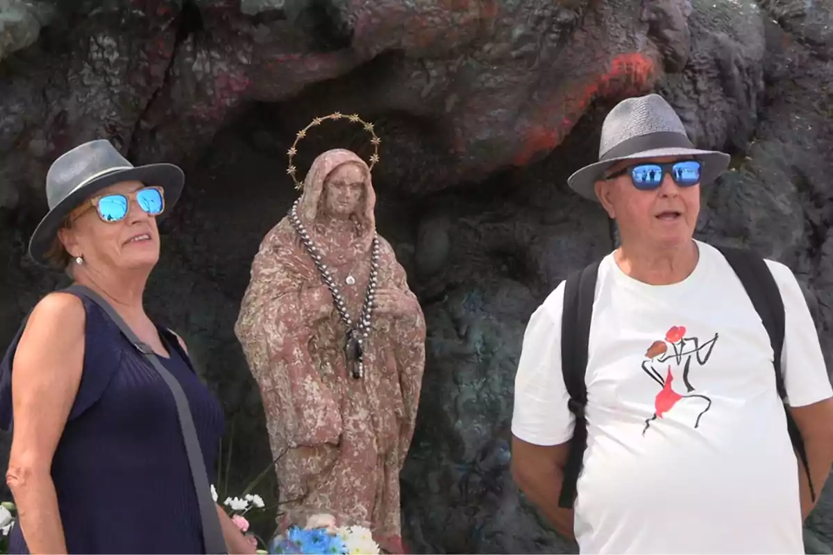 Dos personas mayores con sombreros y gafas de sol posan junto a una estatua religiosa rodeada de flores frente a una pared rocosa Dos personas mayores con sombreros y gafas de sol posan junto a una estatua religiosa rodeada de flores frente a una pared rocosa