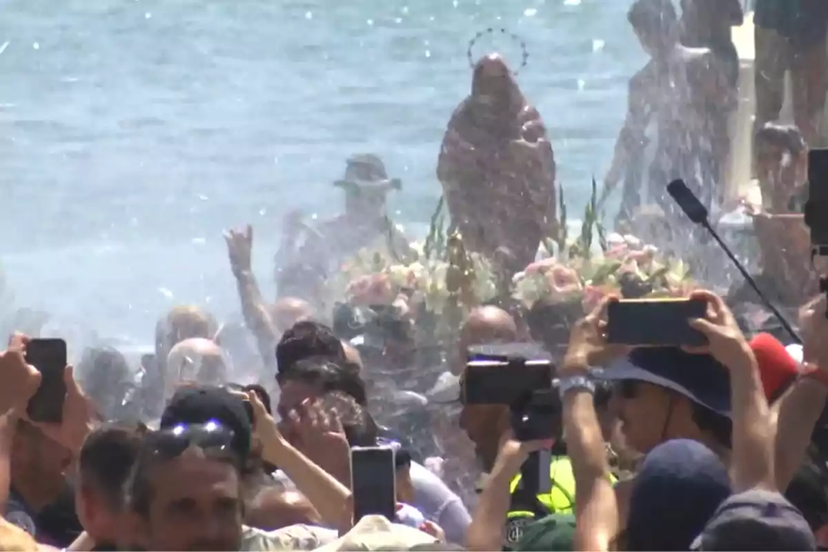 Multitud de personas en la playa rodean una imagen religiosa decorada con flores mientras toman fotos y se mojan con agua. Multitud de personas en la playa rodean una imagen religiosa decorada con flores mientras toman fotos y se mojan con agua.