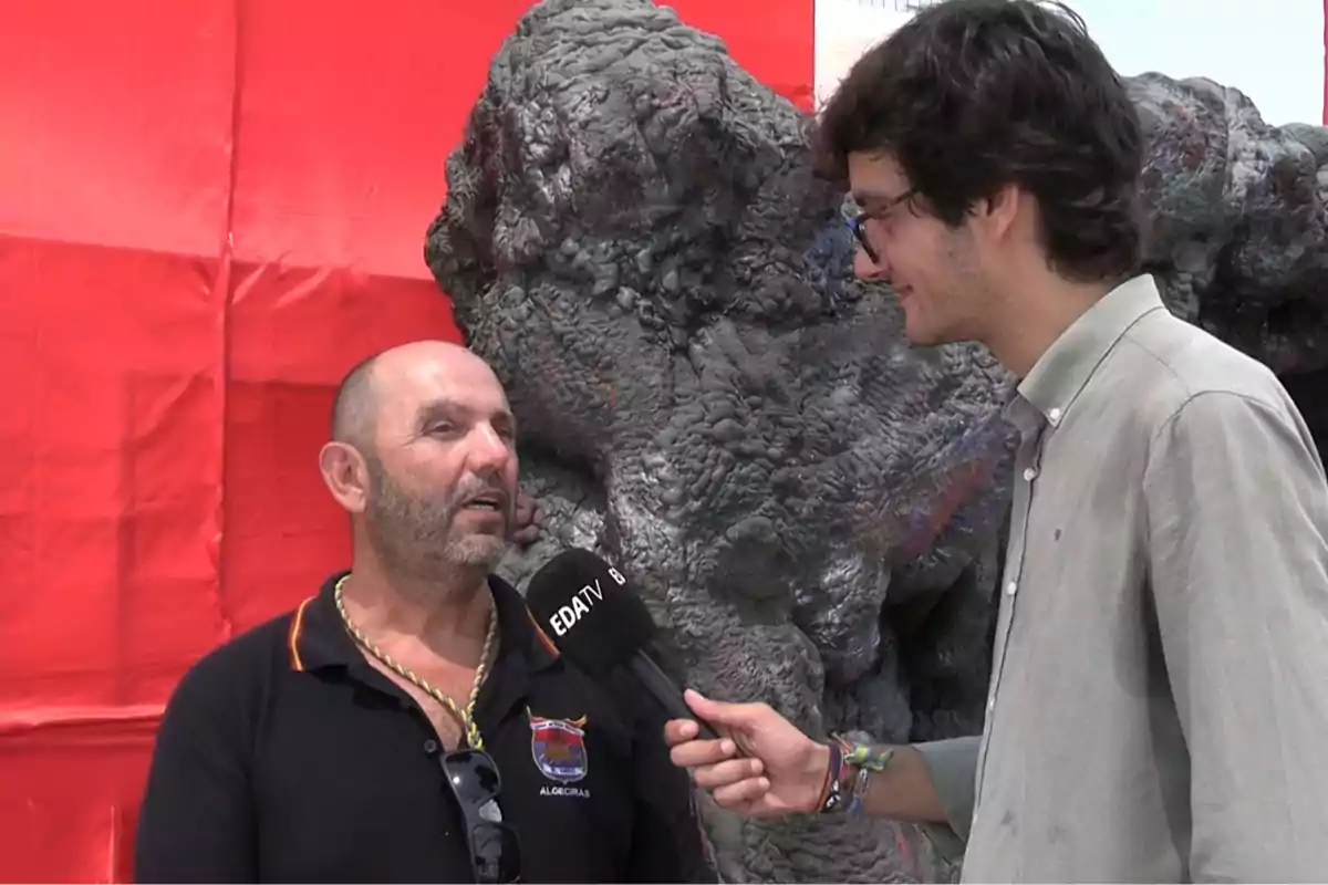 Un hombre con camisa negra y gafas colgadas en el cuello está siendo entrevistado por un joven con camisa gris que sostiene un micrófono frente a un fondo rojo y una estructura gris. Un hombre con camisa negra y gafas colgadas en el cuello está siendo entrevistado por un joven con camisa gris que sostiene un micrófono frente a un fondo rojo y una estructura gris.