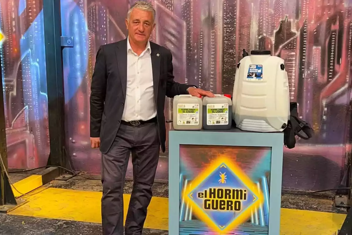 Hombre de traje posando junto a un mostrador con productos y un pulverizador en un set decorado con el logo de El Hormiguero