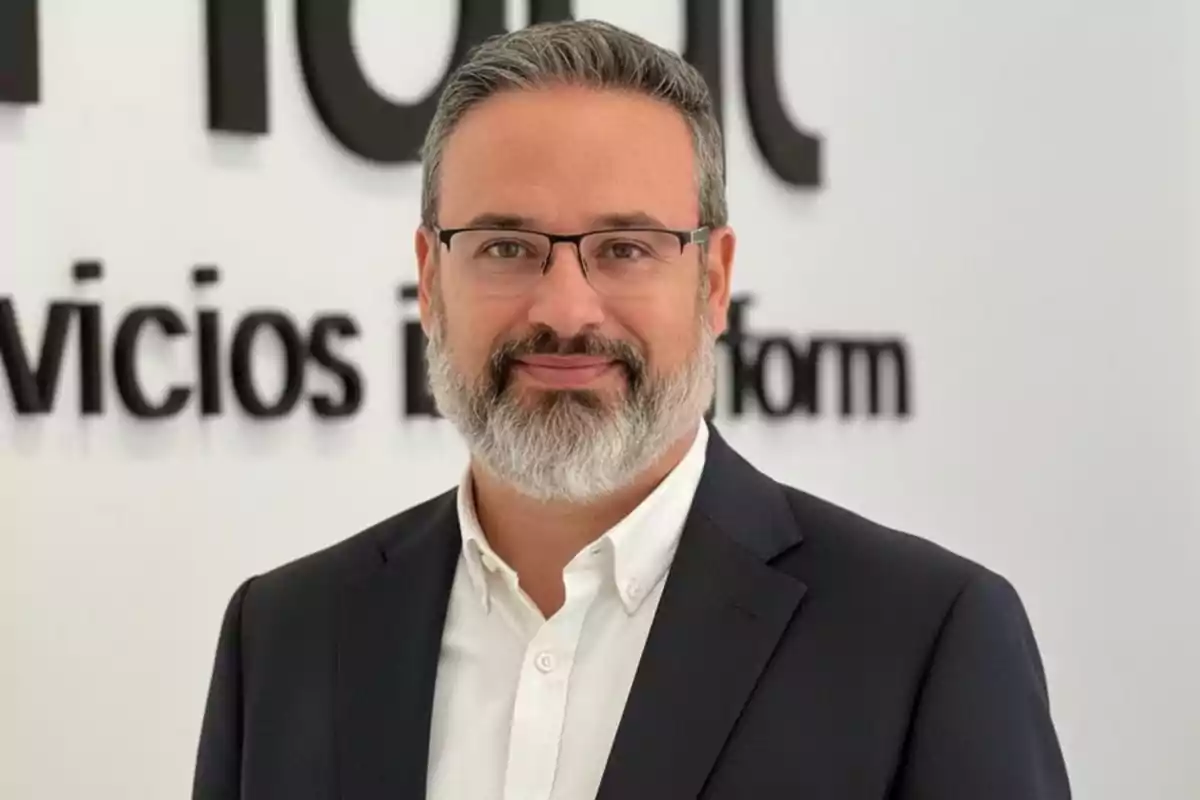 Hombre de cabello y barba canosos con gafas y traje oscuro posando frente a una pared blanca con letras negras.