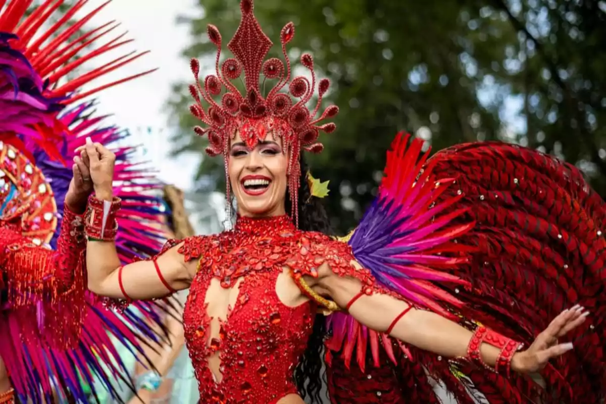 Mujer sonriente con disfraz colorido de plumas rojas y moradas bailando en un desfile o carnaval al aire libre