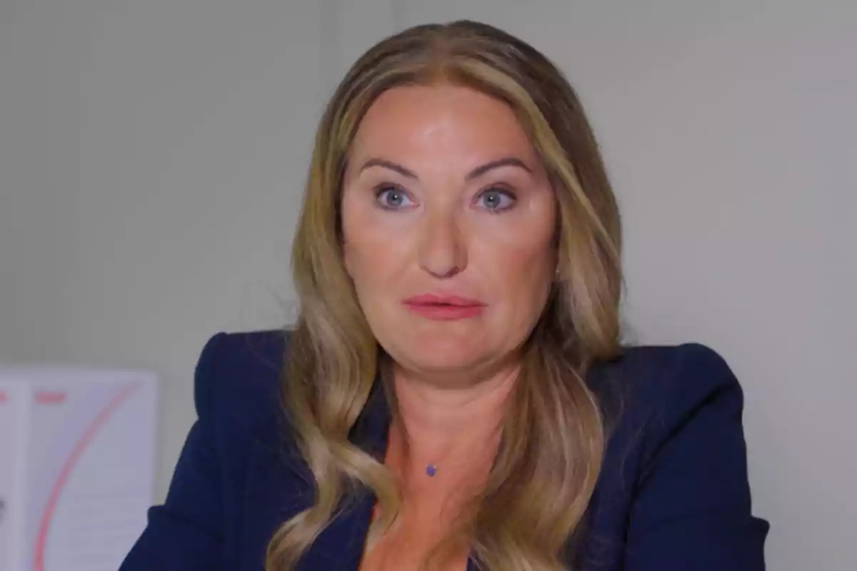 Mujer de cabello largo y rubio con expresión seria, vestida con saco azul oscuro, sentada frente a una pared clara Mujer de cabello largo y rubio con expresión seria, vestida con saco azul oscuro, sentada frente a una pared clara