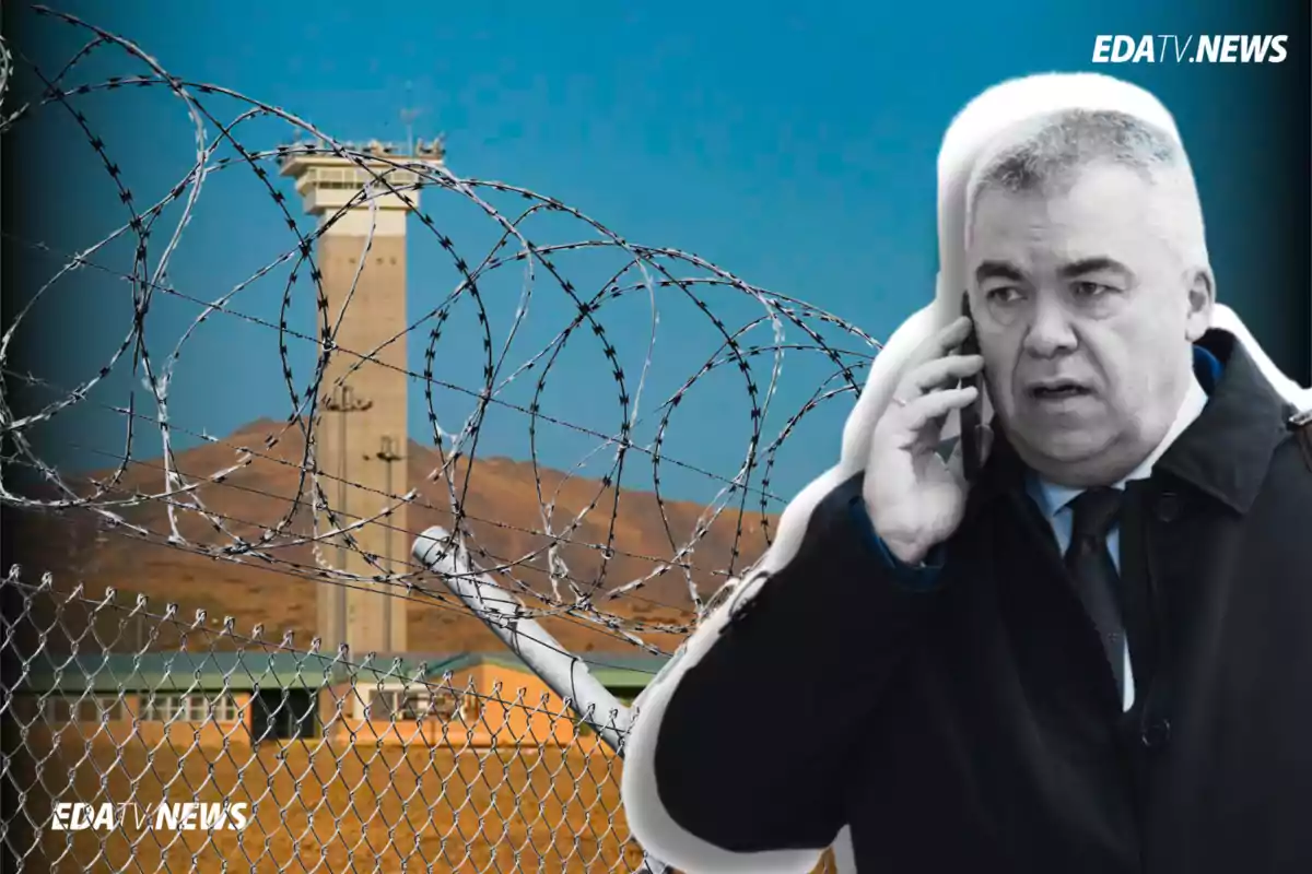 Hombre de traje hablando por teléfono frente a una cerca de alambre de púas con una torre de vigilancia al fondo