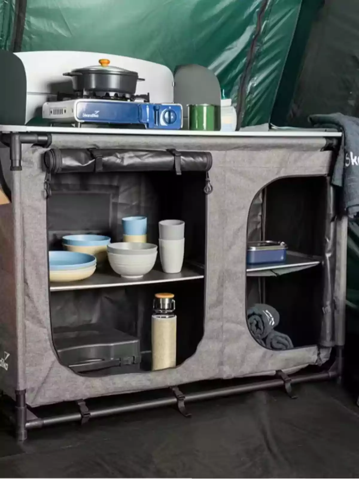 Cocina portátil de camping con estufa, ollas, tazas, platos y utensilios organizados en estantes dentro de una estructura de tela y tubos, ubicada dentro de una tienda de campaña