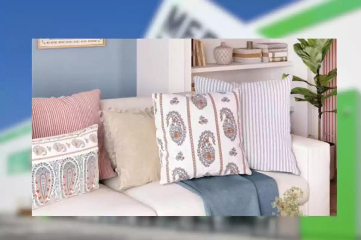 Sofá blanco decorado con cojines de diferentes colores y estampados junto a una manta azul y una planta en el fondo Sofá blanco decorado con cojines de diferentes colores y estampados junto a una manta azul y una planta en el fondo