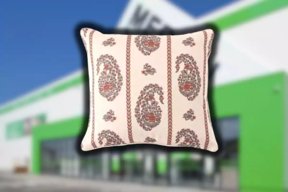 Cojín decorativo con diseño de flores y hojas en tonos marrón y gris sobre fondo claro frente a un edificio desenfocado con detalles verdes y blancos Cojín decorativo con diseño de flores y hojas en tonos marrón y gris sobre fondo claro frente a un edificio desenfocado con detalles verdes y blancos