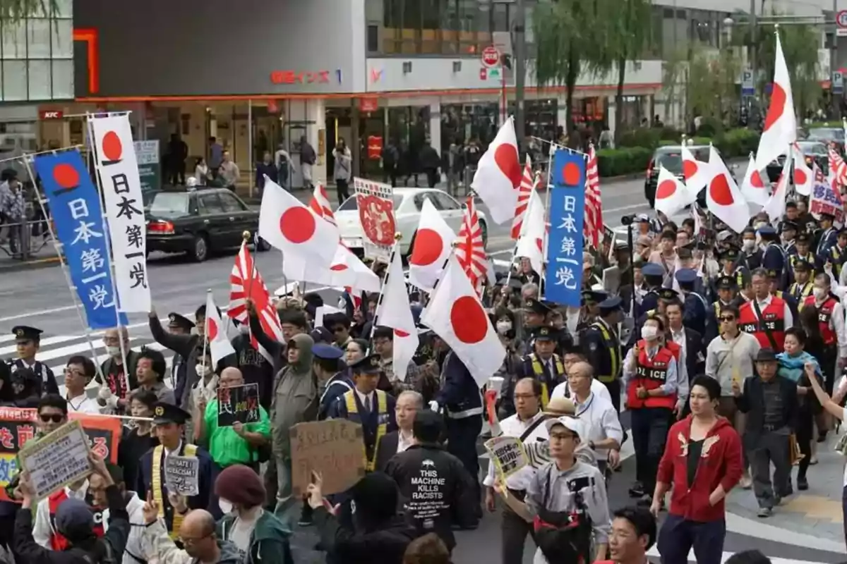 Manifestación en una calle de Japón con numerosas personas portando banderas japonesas y pancartas, acompañadas por policías y observadores Manifestación en una calle de Japón con numerosas personas portando banderas japonesas y pancartas, acompañadas por policías y observadores