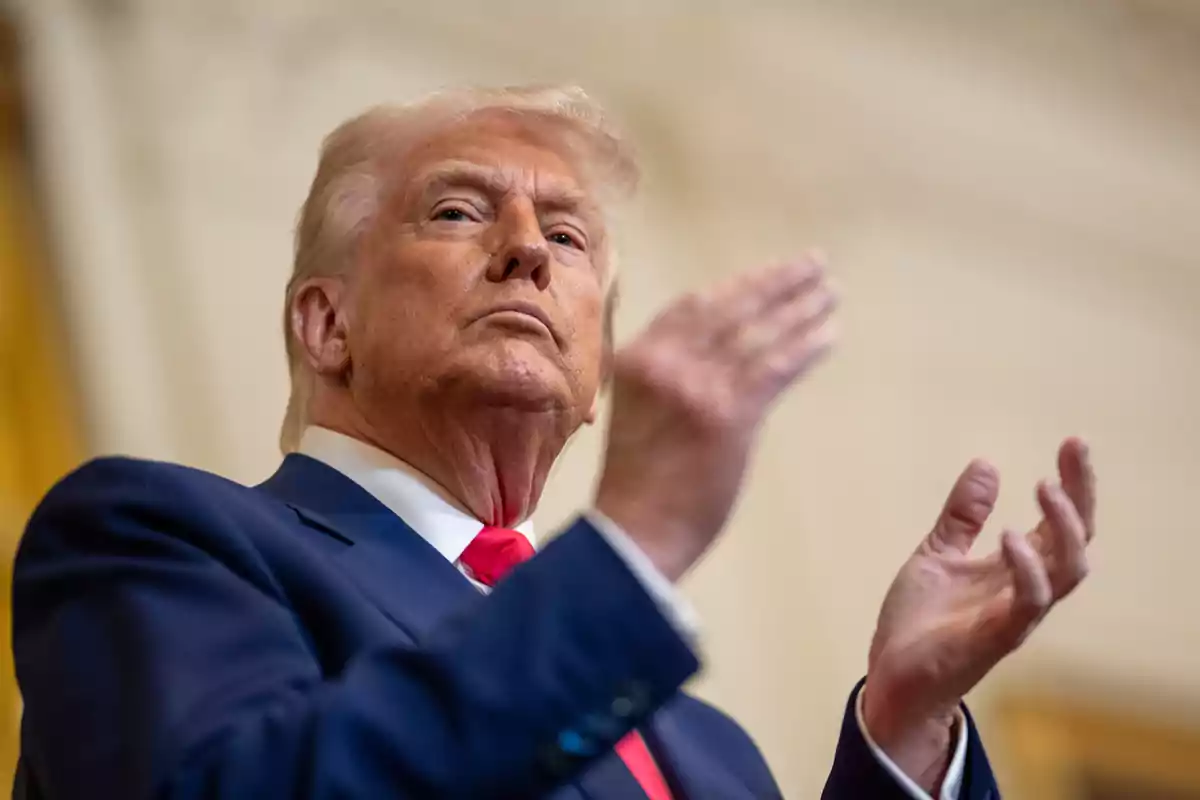 Hombre de cabello rubio con traje azul y corbata roja aplaudiendo en un ambiente interior