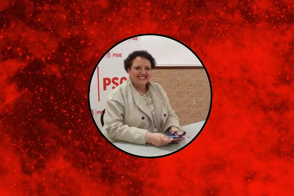 Mujer sonriente sentada en una mesa con fondo de logotipo del PSOE dentro de un círculo sobre un fondo rojo moteado