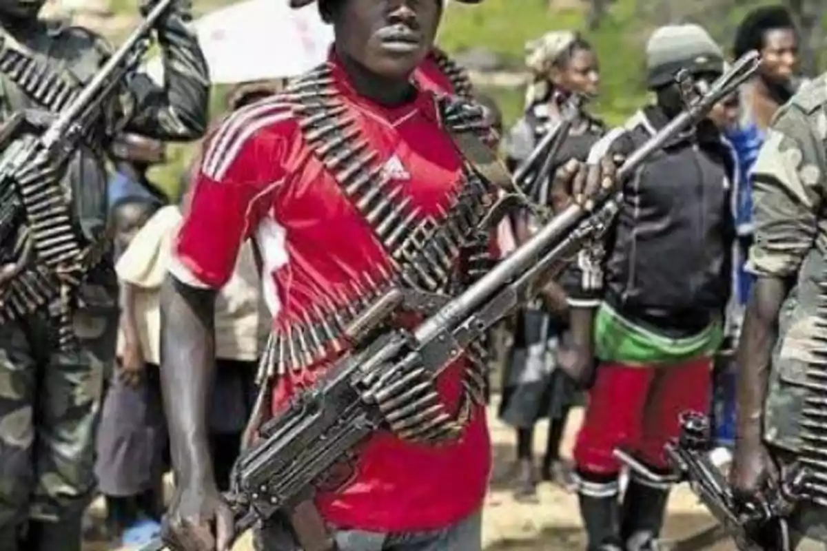 Un grupo de personas armadas con rifles y municiones, una de ellas lleva una camiseta roja y sombrero, están al aire libre en un entorno rural