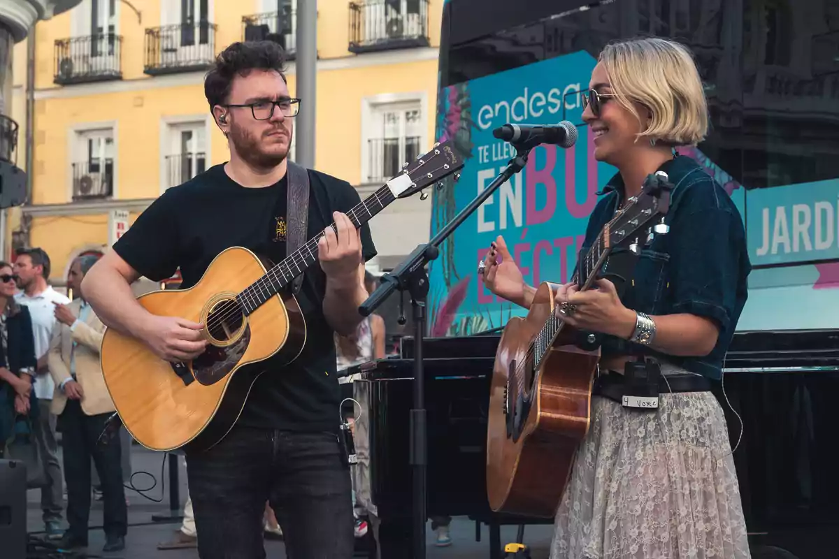 Dos músicos tocan la guitarra y cantan en una presentación al aire libre frente a un grupo de personas