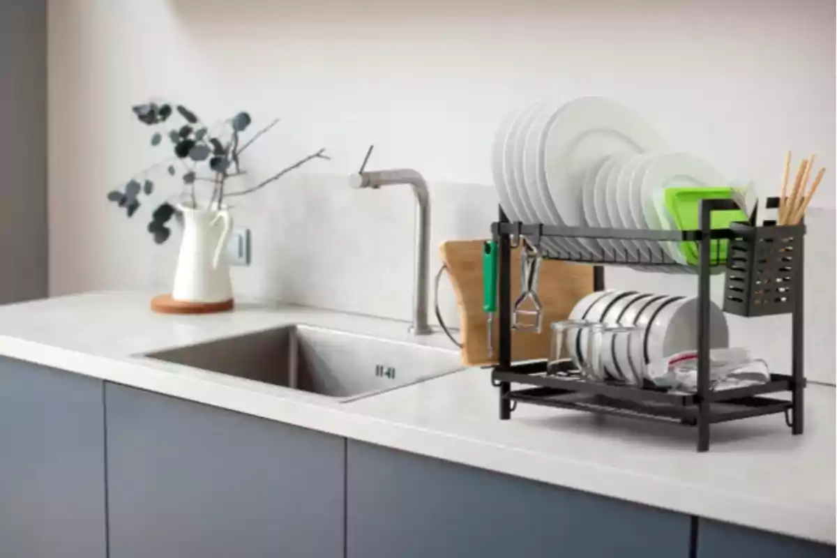 Escurridor de platos negro sobre una encimera blanca junto a un fregadero con platos, vasos y utensilios organizados, al fondo una jarra blanca con ramas decorativas Escurridor de platos negro sobre una encimera blanca junto a un fregadero con platos, vasos y utensilios organizados, al fondo una jarra blanca con ramas decorativas