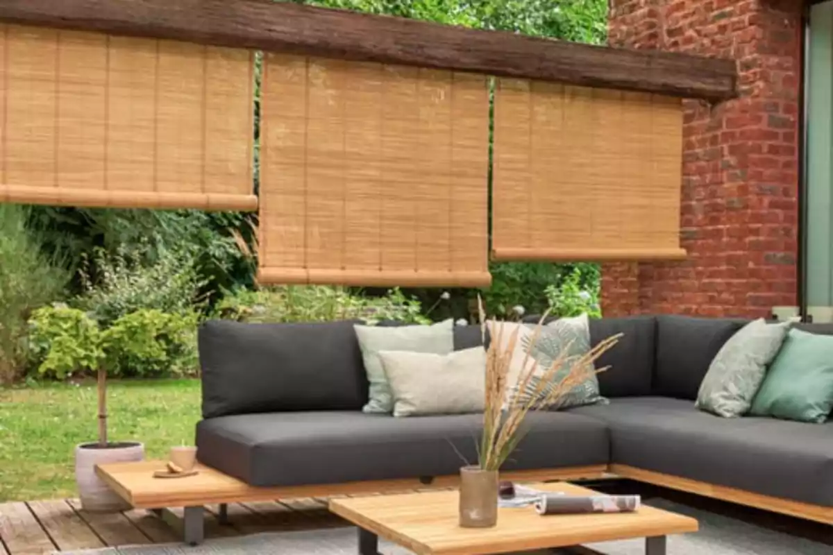 Sala exterior con sofá en L de cojines grises, mesa de centro de madera, persianas enrollables de bambú y plantas alrededor Sala exterior con sofá en L de cojines grises, mesa de centro de madera, persianas enrollables de bambú y plantas alrededor