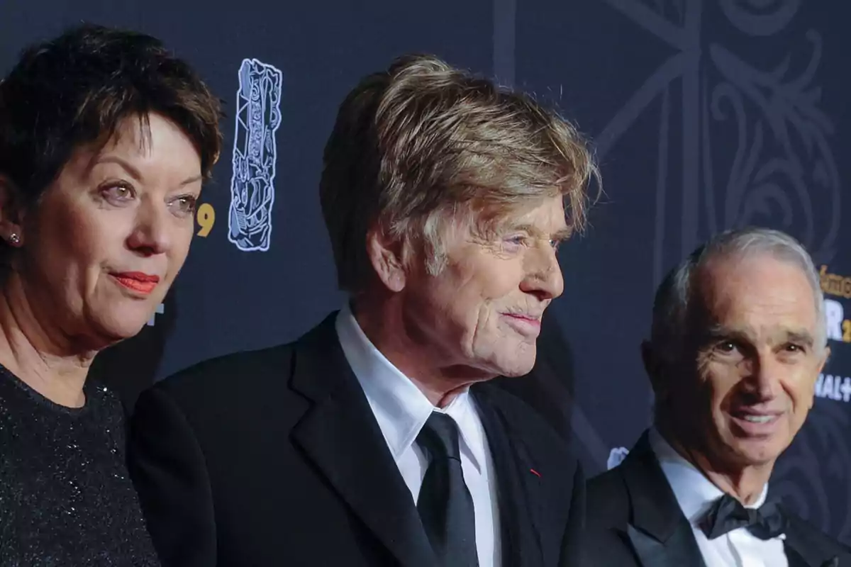 Tres personas vestidas de gala posan en una alfombra roja frente a un fondo oscuro decorado con logotipos y diseños elegantes.