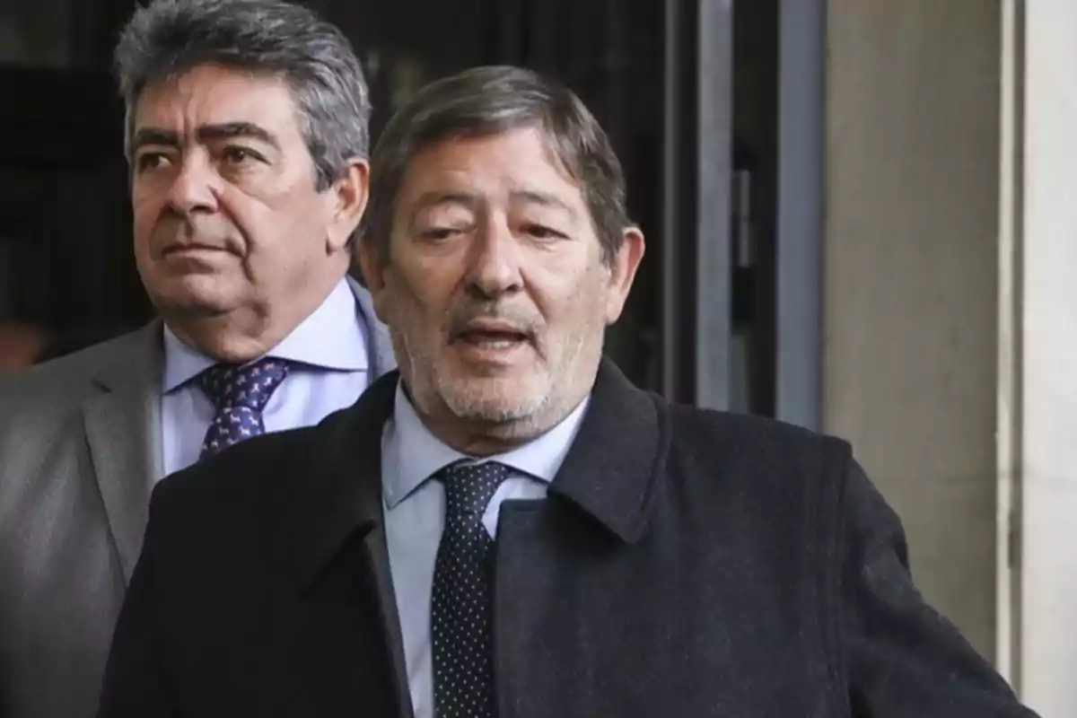 Dos hombres de traje y corbata salen de un edificio, uno de ellos en primer plano con abrigo oscuro y expresión seria.