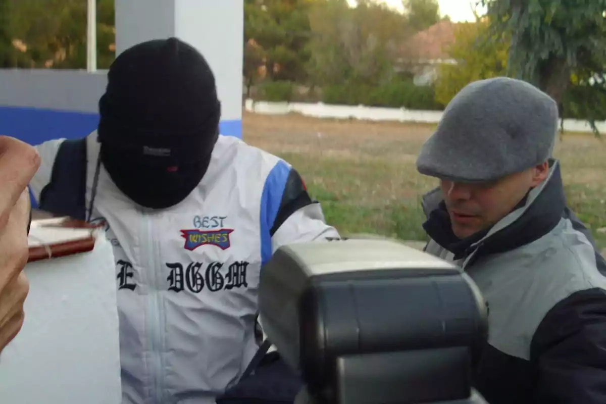 Dos personas con ropa de abrigo y gorros, una de ellas con el rostro cubierto, están al aire libre junto a un objeto grande en primer plano. Dos personas con ropa de abrigo y gorros, una de ellas con el rostro cubierto, están al aire libre junto a un objeto grande en primer plano.