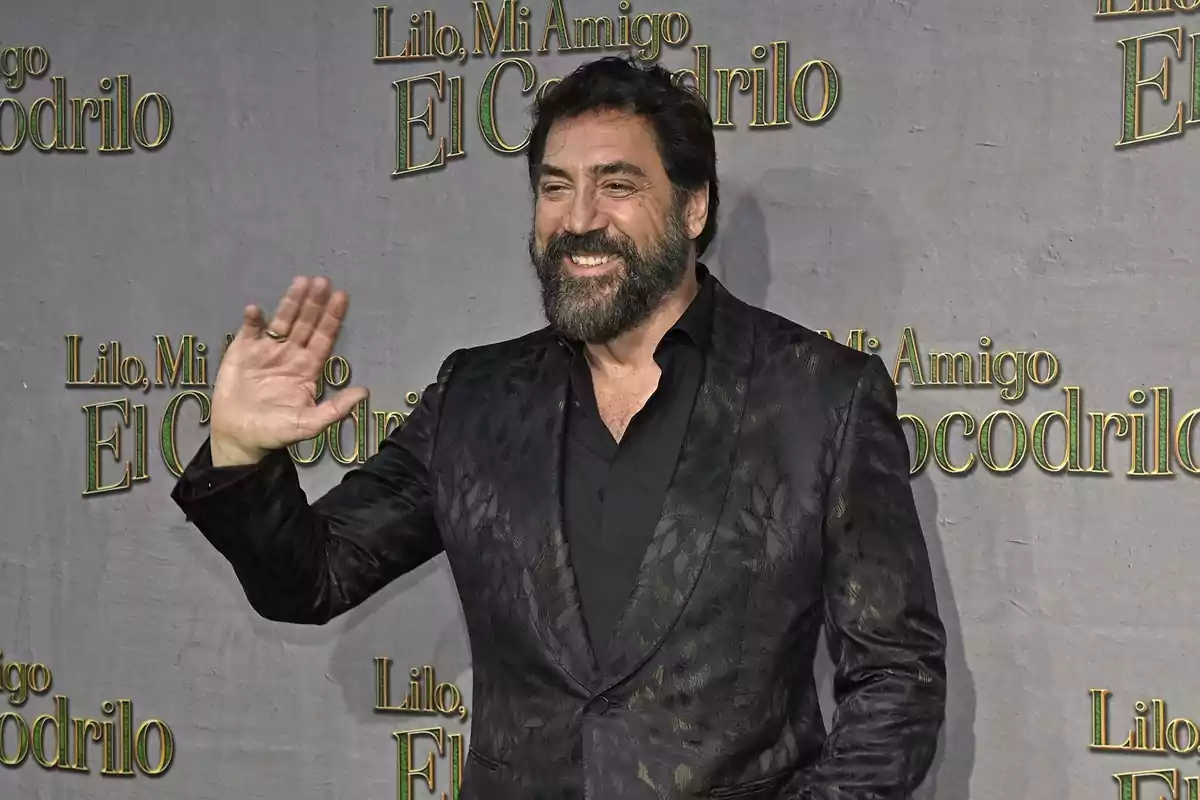 Hombre con barba y cabello oscuro sonríe y saluda con la mano en un evento con fondo promocional de la película Lilo Mi Amigo El Cocodrilo Hombre con barba y cabello oscuro sonríe y saluda con la mano en un evento con fondo promocional de la película Lilo Mi Amigo El Cocodrilo
