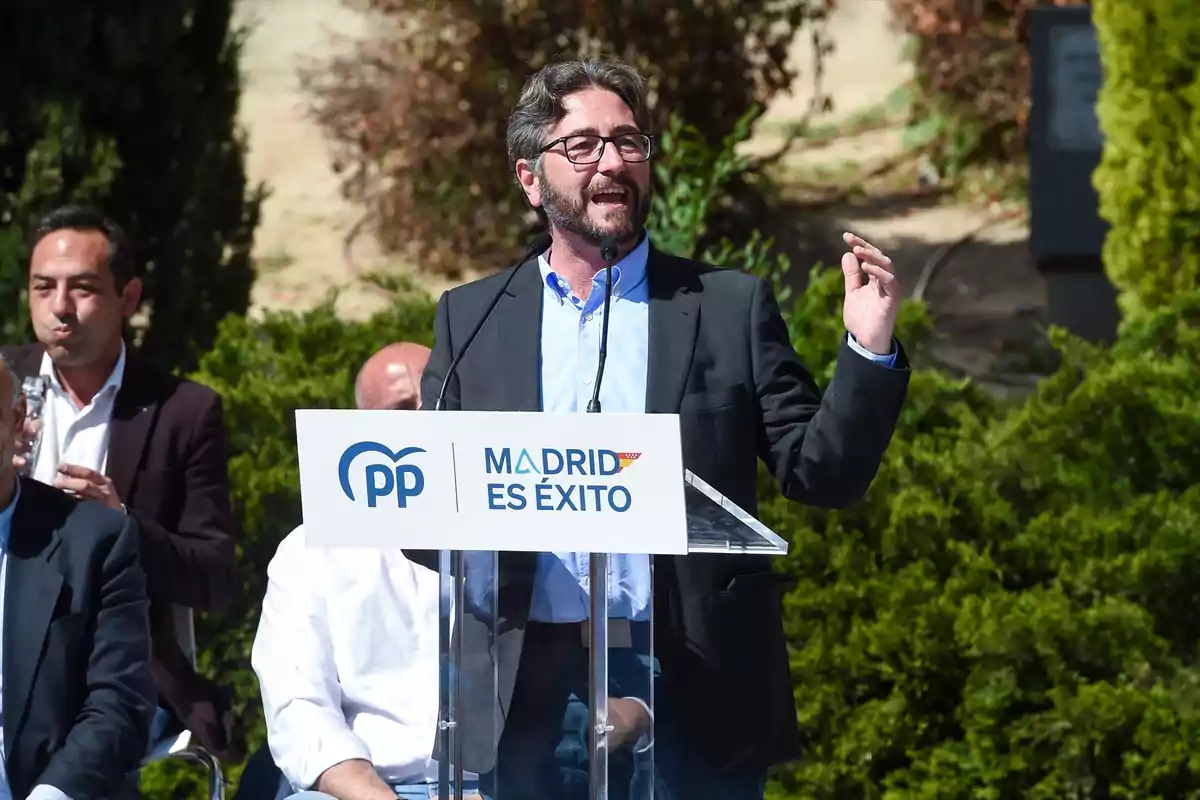 Un hombre con gafas y barba habla en un atril durante un evento al aire libre del Partido Popular con un cartel que dice Madrid es éxito y varias personas sentadas al fondo Un hombre con gafas y barba habla en un atril durante un evento al aire libre del Partido Popular con un cartel que dice Madrid es éxito y varias personas sentadas al fondo
