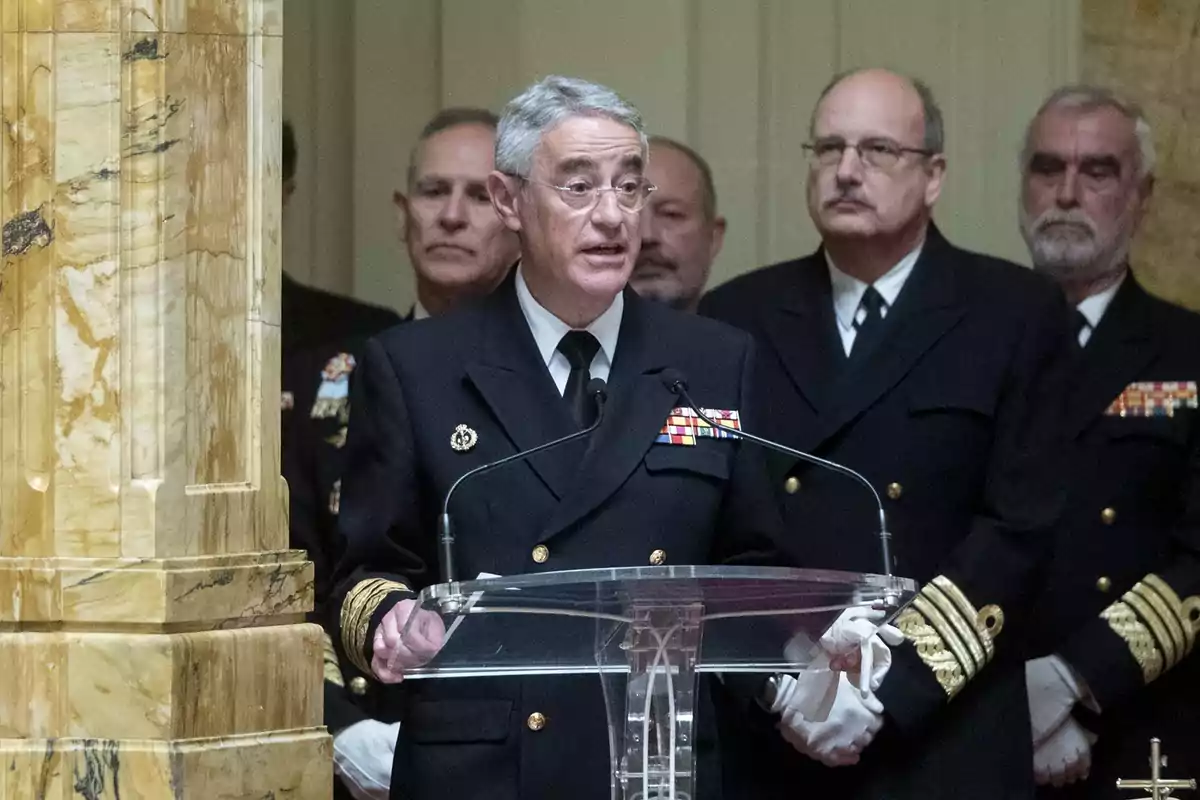 Un grupo de oficiales navales con uniformes formales escucha mientras uno de ellos habla en un atril de cristal en un edificio elegante de mármol.