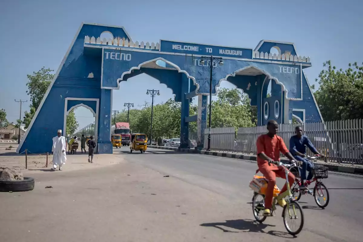 Personas en bicicleta y caminando cerca de un gran arco azul que da la bienvenida a Maiduguri en una calle soleada