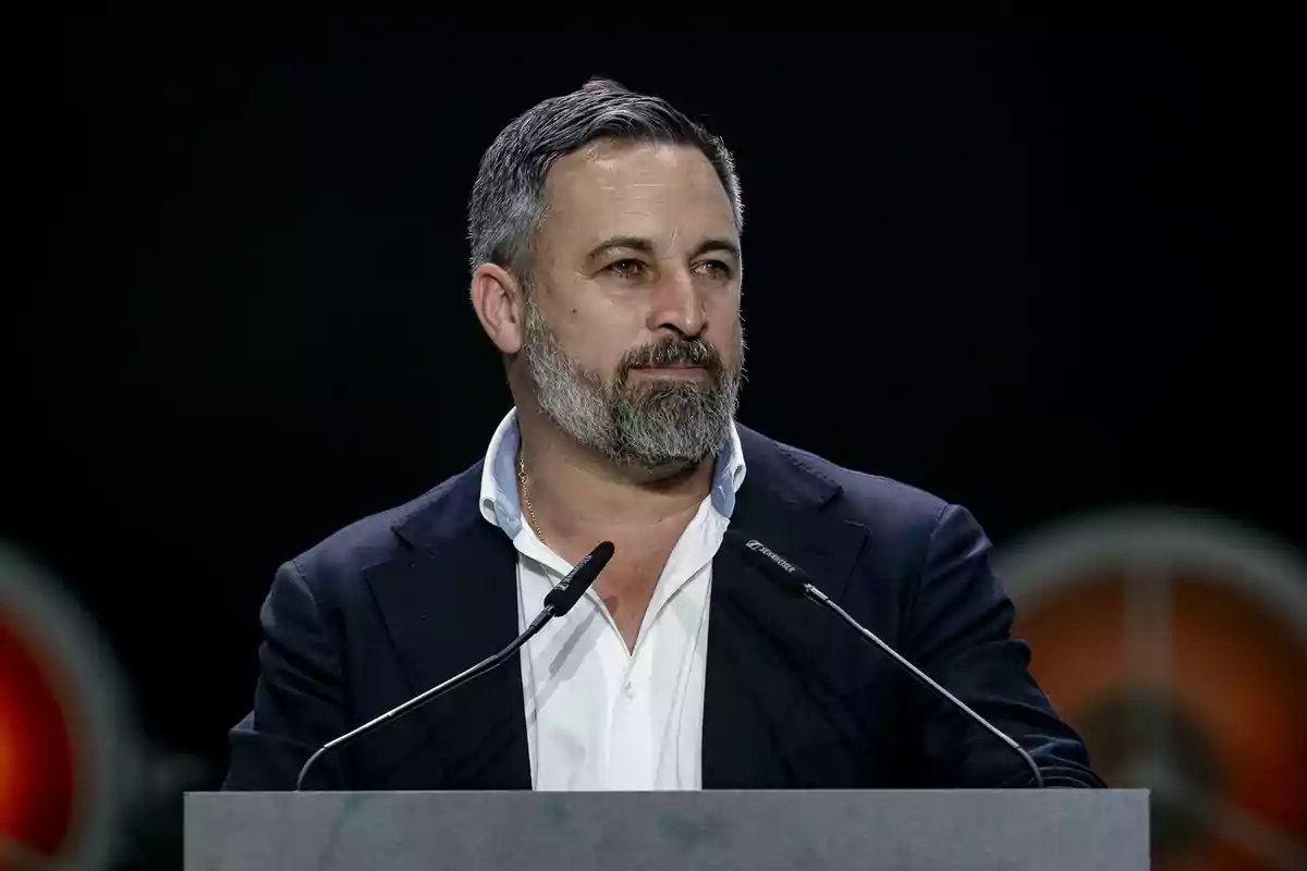 Hombre de barba y cabello corto canoso hablando en un podio con micrófonos y fondo oscuro Hombre de barba y cabello corto canoso hablando en un podio con micrófonos y fondo oscuro