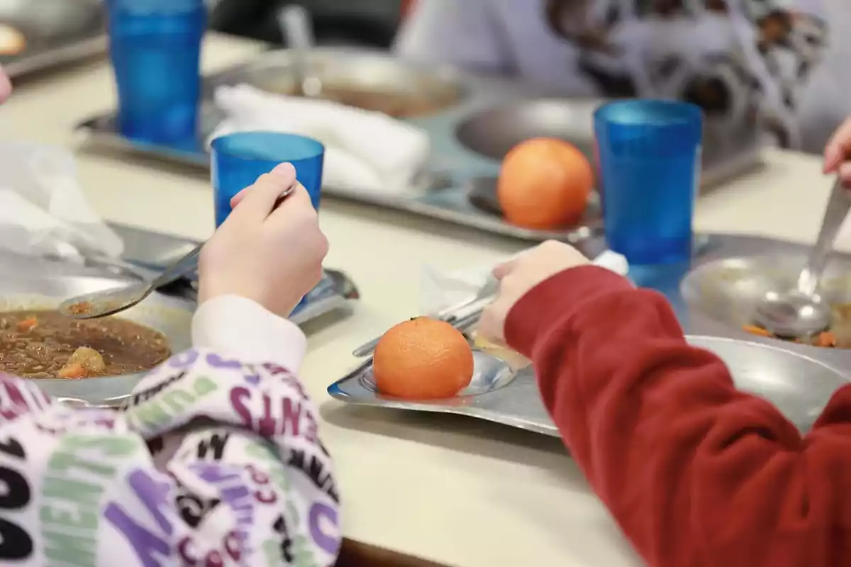 Niños comiendo en un comedor escolar con bandejas de comida, vasos azules y una mandarina Niños comiendo en un comedor escolar con bandejas de comida, vasos azules y una mandarina