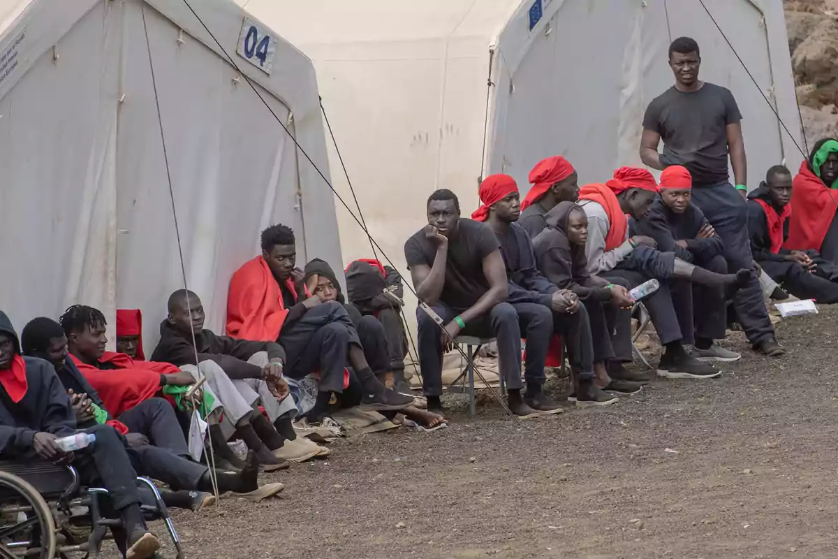 Un grupo de personas sentadas y algunas de pie frente a unas carpas blancas, muchas de ellas llevan mantas o pañuelos rojos y parecen estar esperando en un campamento improvisado sobre suelo de tierra. Un grupo de personas sentadas y algunas de pie frente a unas carpas blancas, muchas de ellas llevan mantas o pañuelos rojos y parecen estar esperando en un campamento improvisado sobre suelo de tierra.