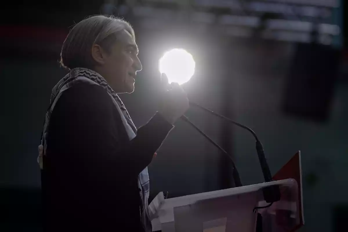 Persona de cabello canoso hablando en un podio con micrófonos y una luz brillante detrás de su cabeza Persona de cabello canoso hablando en un podio con micrófonos y una luz brillante detrás de su cabeza