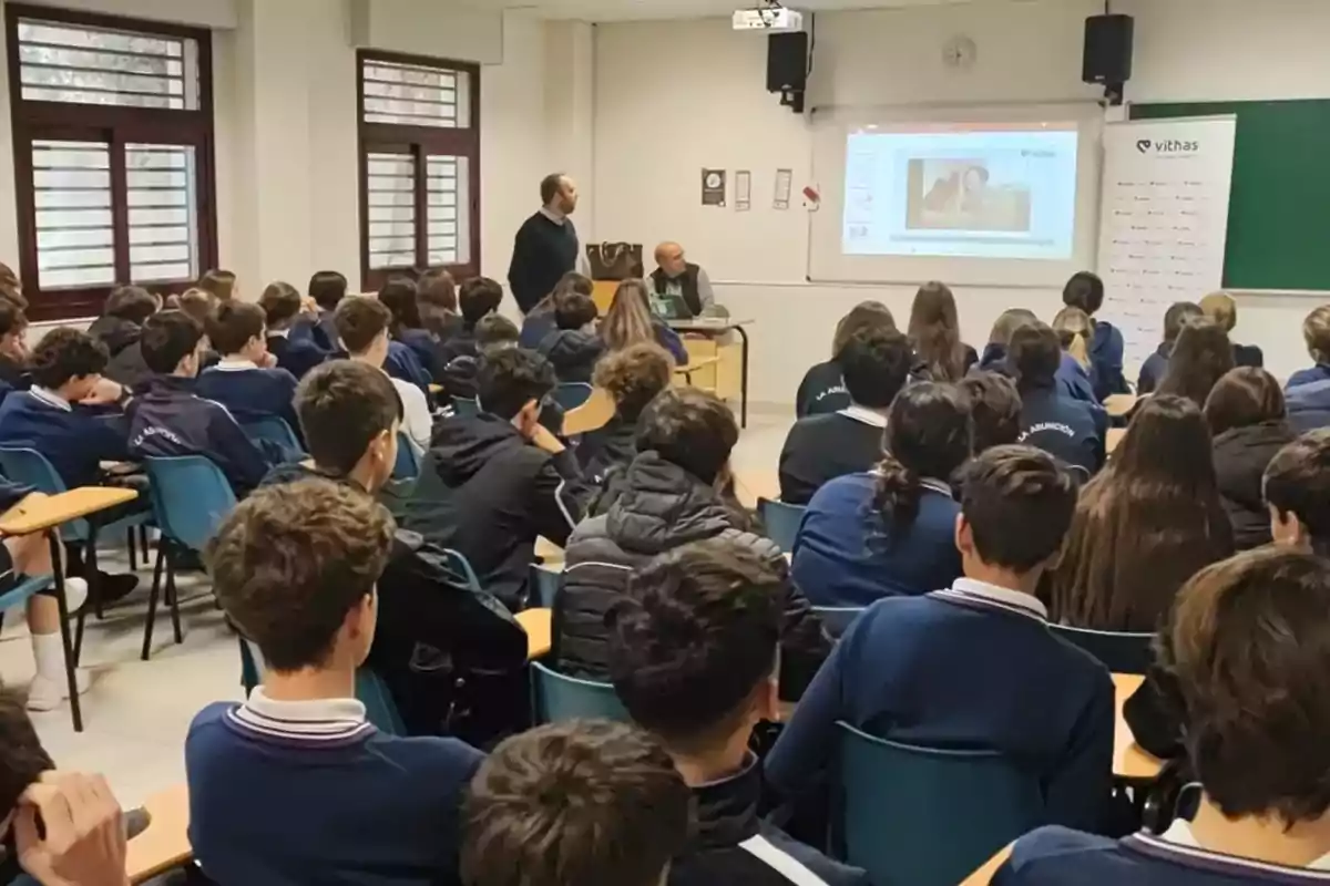 Un grupo de estudiantes en un aula atiende una presentación proyectada en la pared mientras dos adultos están al frente de la clase