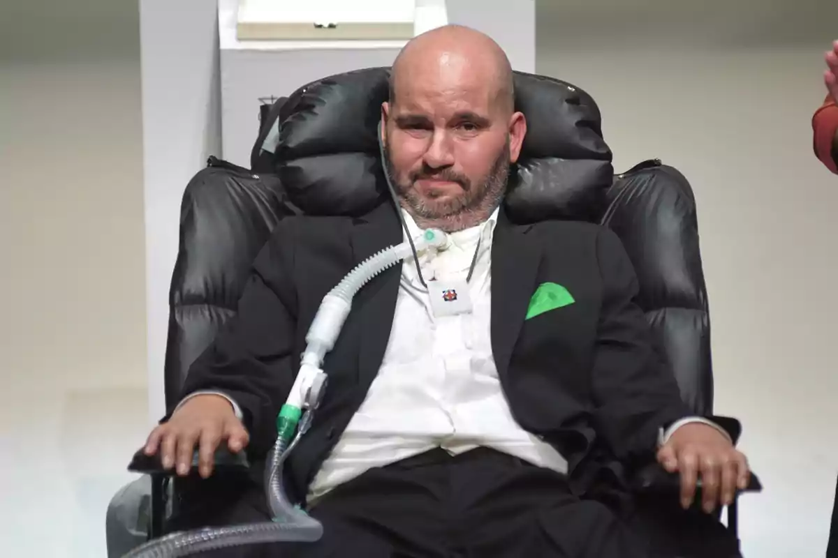 Hombre calvo con barba sentado en una silla de ruedas eléctrica usando un respirador y vestido con traje negro y pañuelo verde en el bolsillo Hombre calvo con barba sentado en una silla de ruedas eléctrica usando un respirador y vestido con traje negro y pañuelo verde en el bolsillo