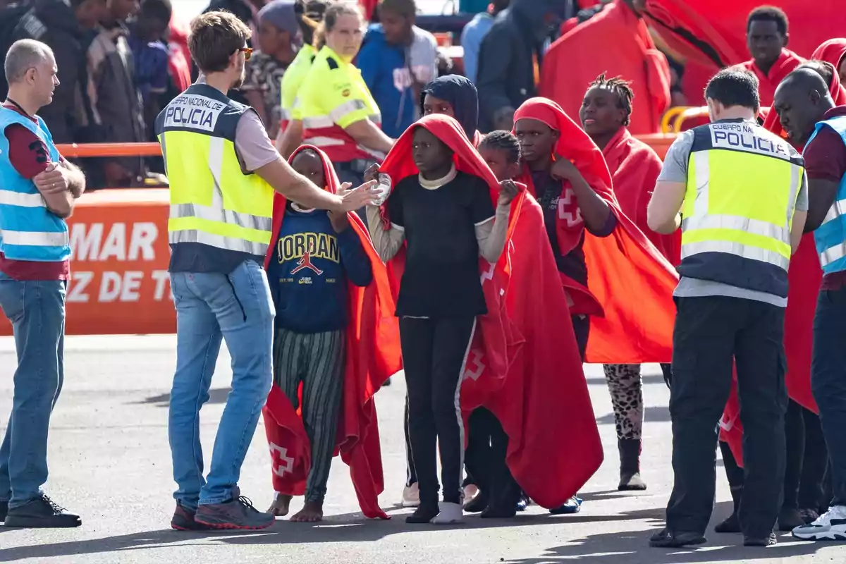Un grupo de personas migrantes cubiertas con mantas rojas es atendido por policías y personal de emergencias en una zona exterior Un grupo de personas migrantes cubiertas con mantas rojas es atendido por policías y personal de emergencias en una zona exterior