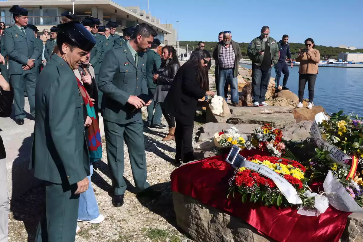 Un grupo de personas, incluyendo varios agentes uniformados, rinde homenaje junto a una ofrenda floral colocada sobre una roca cerca del agua en un día soleado.