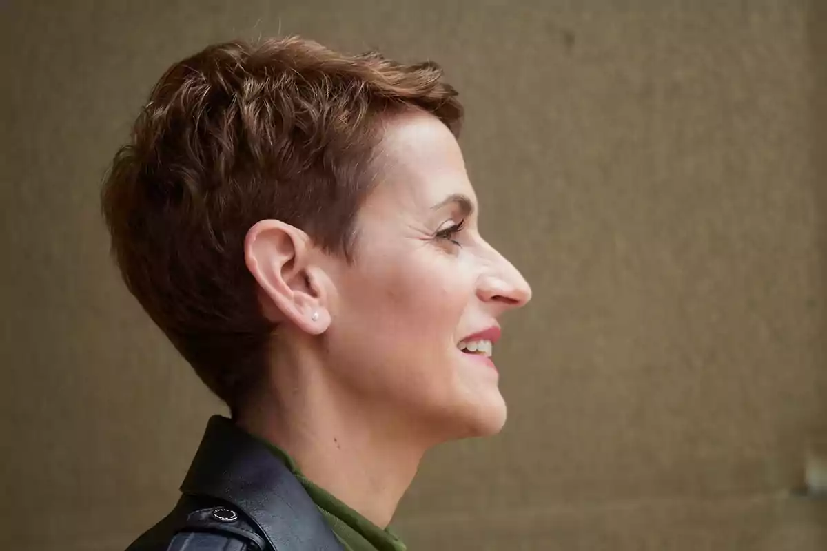 Mujer de perfil con cabello corto castaño y chaqueta negra sonriendo frente a un fondo beige Mujer de perfil con cabello corto castaño y chaqueta negra sonriendo frente a un fondo beige
