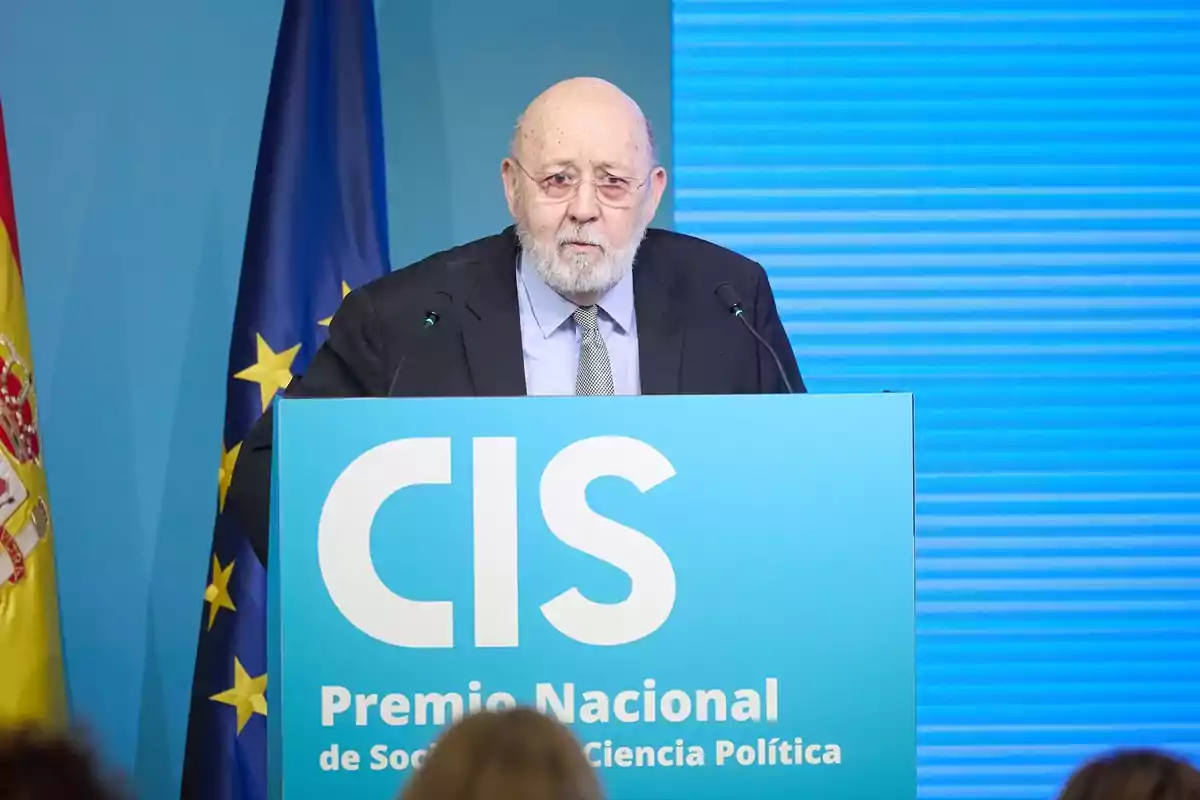 Hombre mayor con barba y gafas hablando en un podio azul del CIS durante un evento de premiación con banderas de España y la Unión Europea al fondo