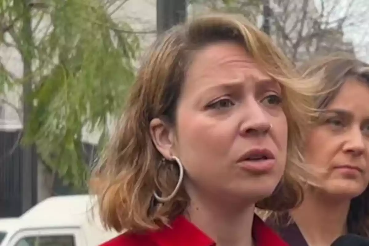 Mujer de cabello rubio corto con expresión seria hablando al aire libre acompañada de otra mujer en segundo plano Mujer de cabello rubio corto con expresión seria hablando al aire libre acompañada de otra mujer en segundo plano
