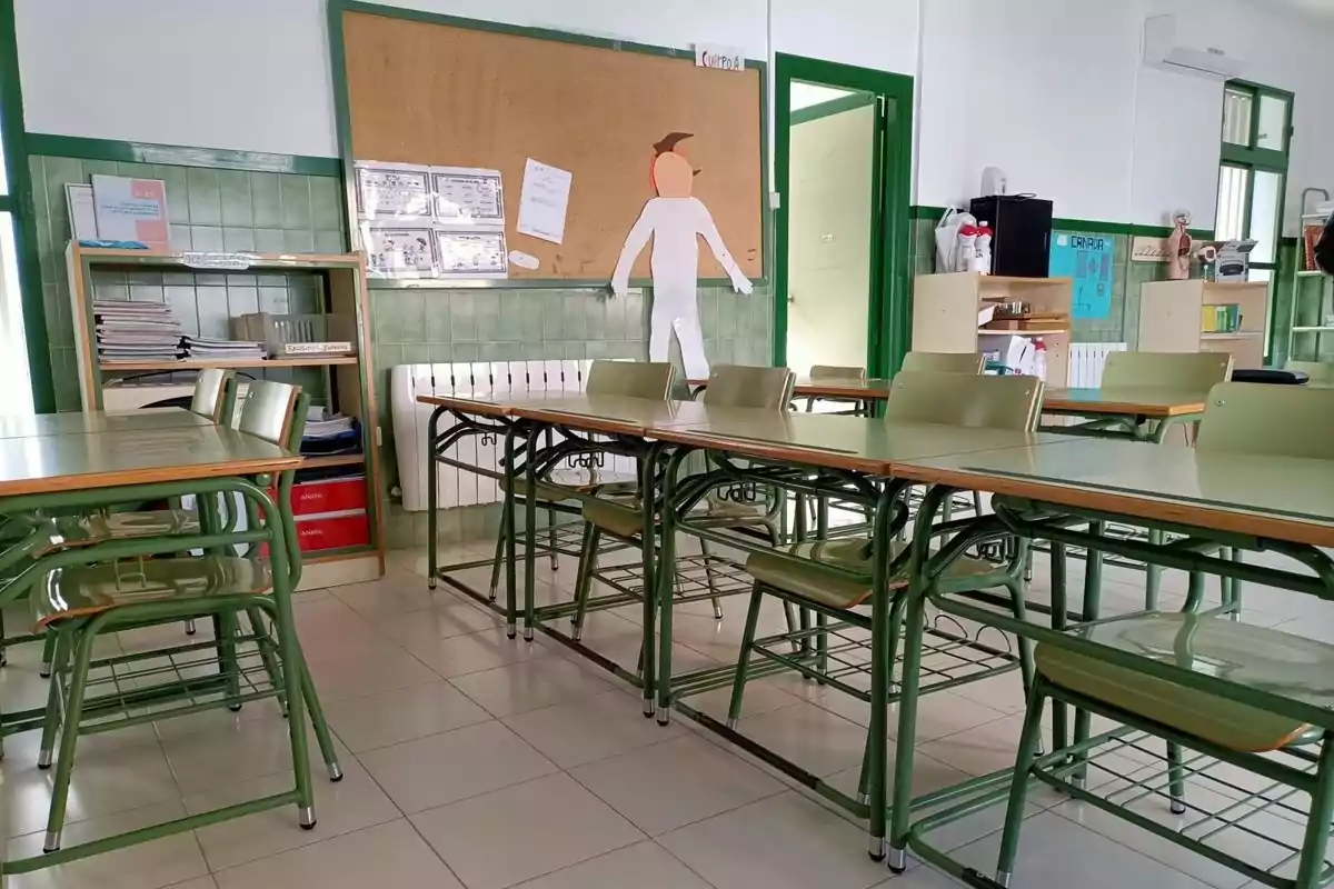 Aula escolar vacía con pupitres y sillas verdes, estantería con libros y papeles, una figura de papel en la pared y materiales educativos en el fondo