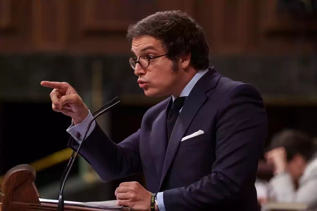 Hombre con gafas y traje oscuro señalando con el dedo mientras habla en un podio Hombre con gafas y traje oscuro señalando con el dedo mientras habla en un podio