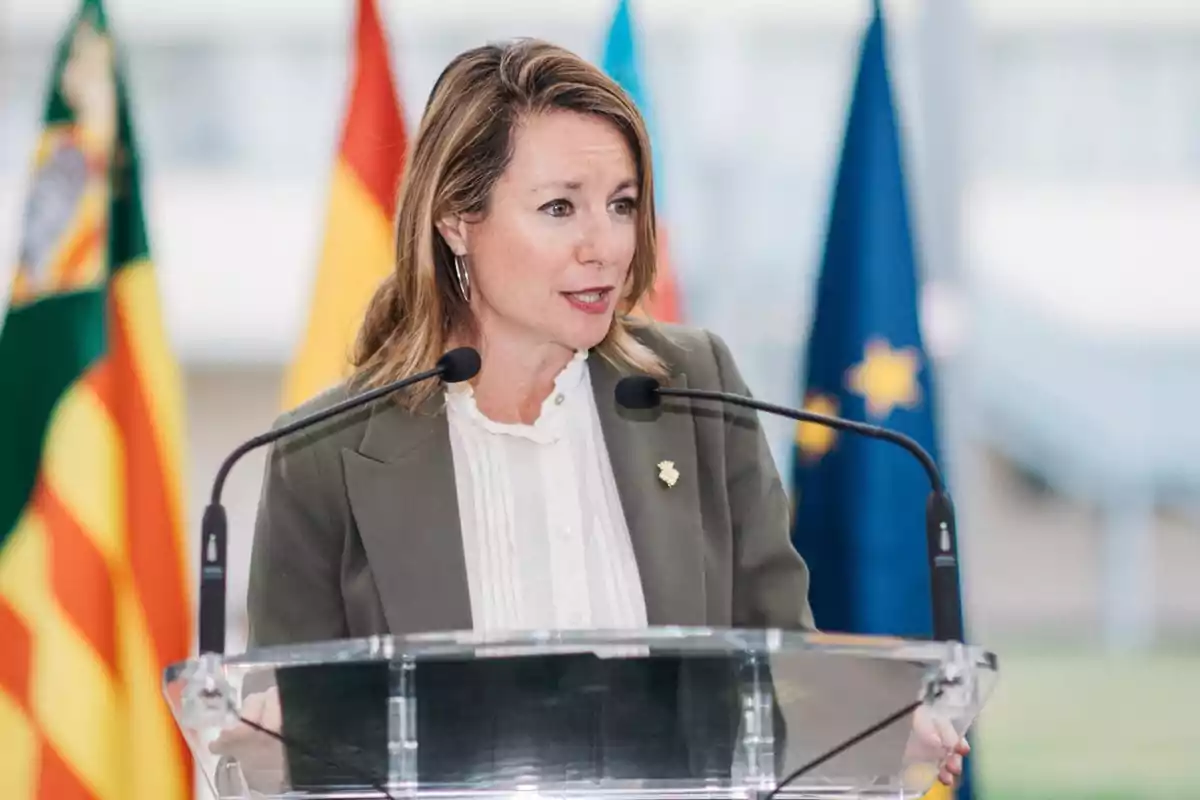 Mujer hablando en un podio con micrófonos y varias banderas de fondo